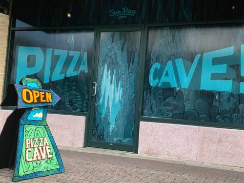 A storefront with "Pizza Cave" painted on the windows and an open sign in front, featuring a cave-themed design.