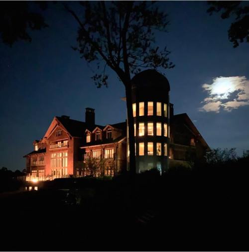 A large, illuminated house at night, with a moonlit sky and trees in the foreground.