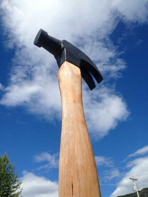 A large wooden hammer sculpture against a blue sky with clouds.