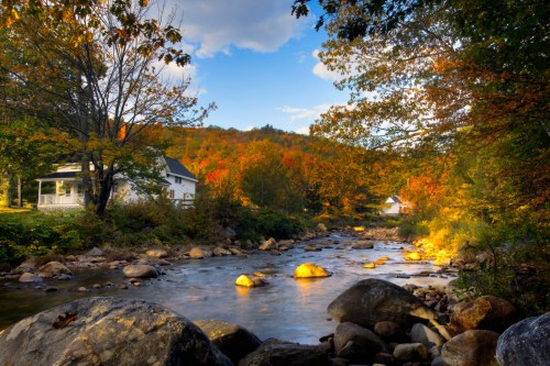A serene river flows past a house, surrounded by vibrant autumn foliage and rocky banks under a blue sky.