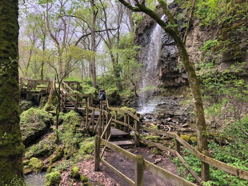 A person walks along a wooden path beside a waterfall in a lush, green forest.