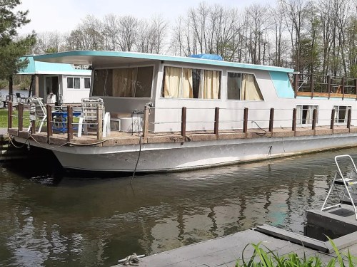 A houseboat docked on calm water, surrounded by trees, with a deck and outdoor seating visible.