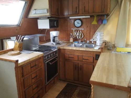 Cozy kitchen in a boat with wooden cabinets, a stove, sink, and coffee maker, featuring nautical decor and warm lighting.