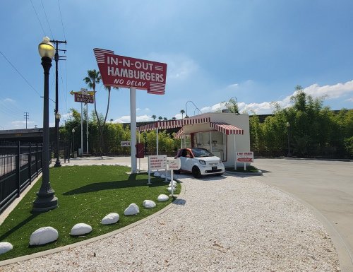 A small In-N-Out Burger stand with a white car parked outside, surrounded by greenery and a clear blue sky.
