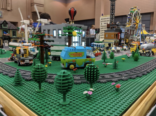 A colorful LEGO city scene with buildings, vehicles, trees, and a train track, featuring a Mystery Machine van.