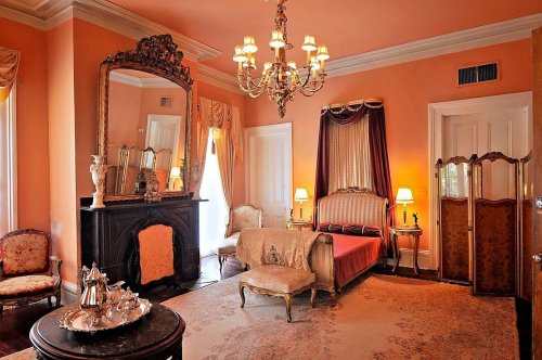 Elegant bedroom with orange walls, vintage furniture, a chandelier, and a large mirror, creating a warm, inviting atmosphere.