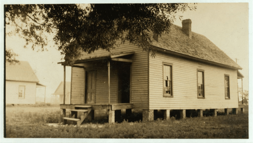 A vintage photograph of a wooden house on stilts, featuring a porch and several windows, surrounded by grass.