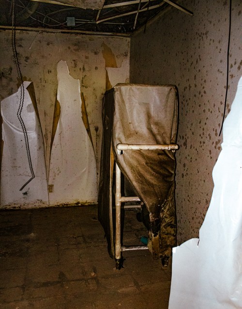 A dimly lit room with a covered cart and peeling walls, featuring shadowy figures outlined on the surface.