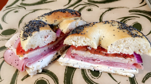 A sliced bagel filled with layers of turkey, ham, cream cheese, tomatoes, and red onions, topped with poppy seeds.