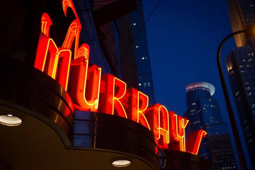 Neon sign reading "Murray" illuminated at night, with city buildings in the background.