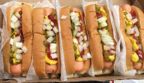 A box of five hot dogs topped with mustard, ketchup, onions, and pickles, served in soft buns.