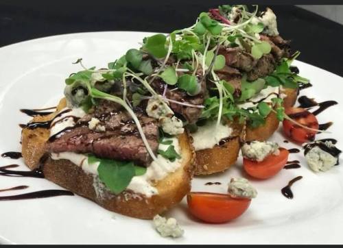 A plate of toasted bread topped with steak, microgreens, blue cheese, and cherry tomatoes, drizzled with balsamic glaze.
