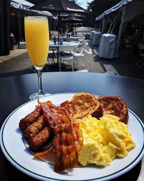 A plate of breakfast with scrambled eggs, bacon, sausage, and French toast, alongside a mimosa on a table outdoors.