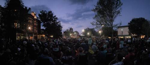 A large crowd gathers in a park at dusk, with trees and buildings illuminated by soft lights.