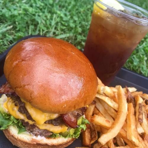 A juicy cheeseburger with bacon, lettuce, and a golden bun, served with crispy fries and a drink on a grassy background.