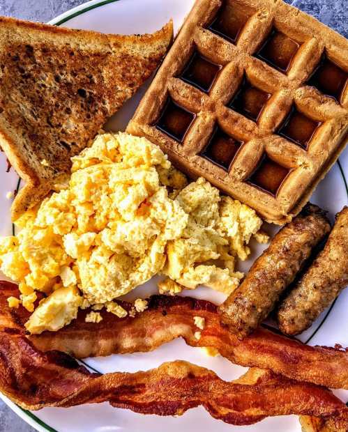 A plate of breakfast featuring scrambled eggs, waffles, toast, sausage links, and crispy bacon.
