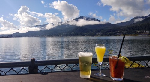 Three drinks on a table with a scenic view of mountains and a lake under a partly cloudy sky.