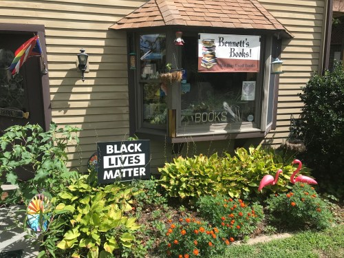 A cozy bookstore with a sign reading "Bennett's Books!" and a "Black Lives Matter" sign in a vibrant garden.