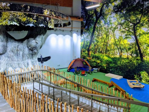 A nature-themed play area featuring a large tree, a tent, and a vibrant forest mural on the wall.