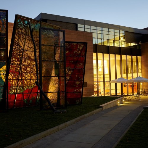 Modern building with large glass windows and colorful stained glass panels, surrounded by a landscaped area at dusk.
