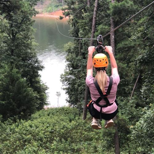 A person zip-lining over a forested area with a lake in the background, wearing a helmet and harness.