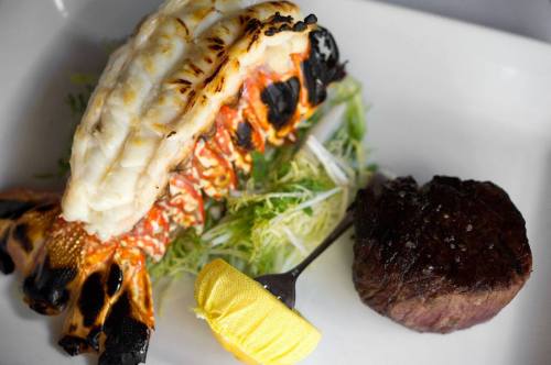 Grilled lobster tail and a tender beef filet served on a bed of greens with a lemon wedge.