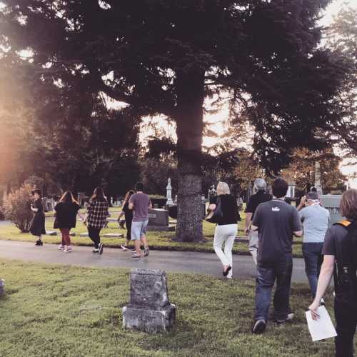 A group of people walking through a cemetery, surrounded by trees and gravestones, during sunset.