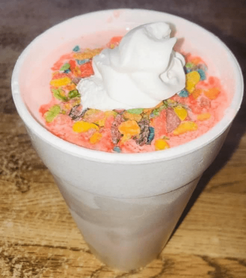 A frosty pink drink topped with whipped cream and colorful cereal pieces in a white cup.