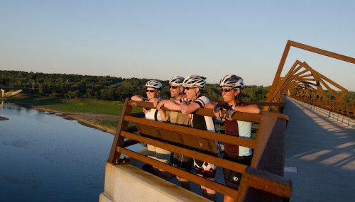 Four cyclists in helmets lean on a bridge railing, enjoying a scenic view of a river and green landscape at sunset.