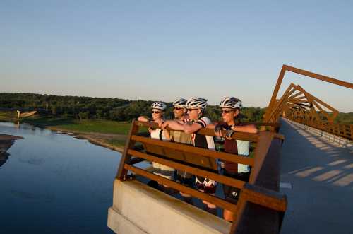 Four cyclists in helmets lean on a bridge railing, overlooking a river and green landscape at sunset.