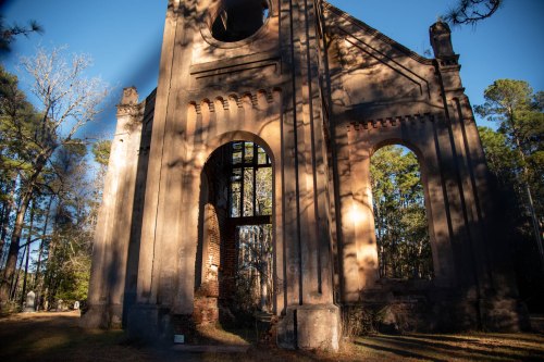 Ruins of a historic building surrounded by trees, with sunlight casting shadows on the weathered walls.