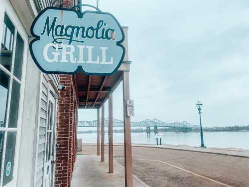 Sign for Magnolia Grill with a view of a river and bridge in the background, under a cloudy sky.