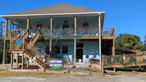 A two-story building with a light blue exterior, wooden stairs, and a sign for a restaurant. Surrounded by greenery.
