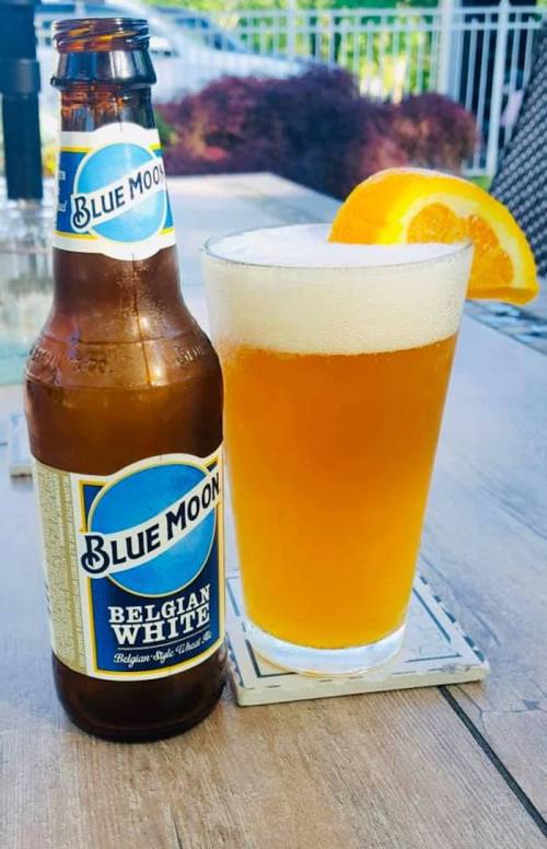 A bottle of Blue Moon Belgian White beer next to a glass of the same beer, garnished with an orange slice.