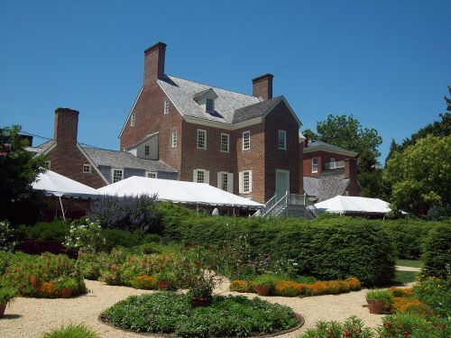 A historic brick house surrounded by lush gardens and white tents under a clear blue sky.