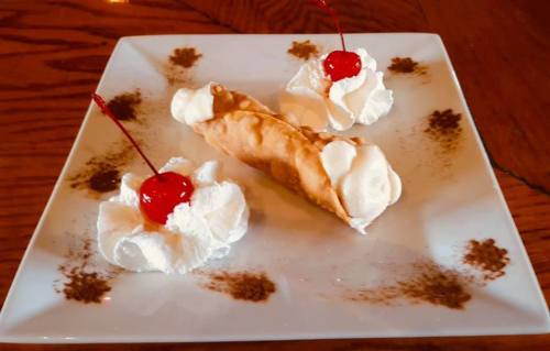 A dessert plate featuring a cannoli with whipped cream and cherries, garnished with chocolate powder.
