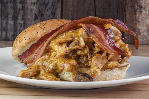 A hearty sandwich with pulled pork, melted cheese, and crispy bacon on a poppy seed bun, served on a plate.