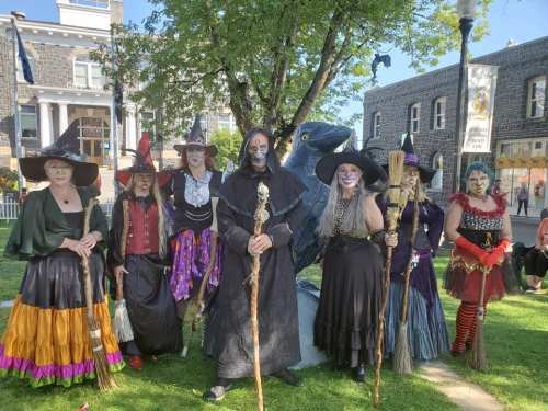 A group of people in witch costumes poses together outdoors, surrounded by trees and buildings.