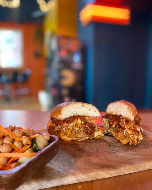 A close-up of a pulled chicken sandwich with sauce and shredded carrots, served with a side of mixed beans on a wooden board.
