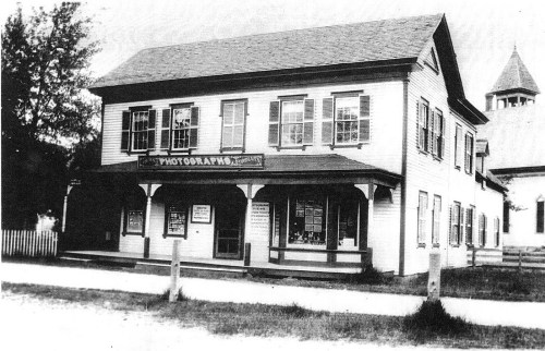 Historic black-and-white photo of a two-story building with a sign for a photography studio and large windows.