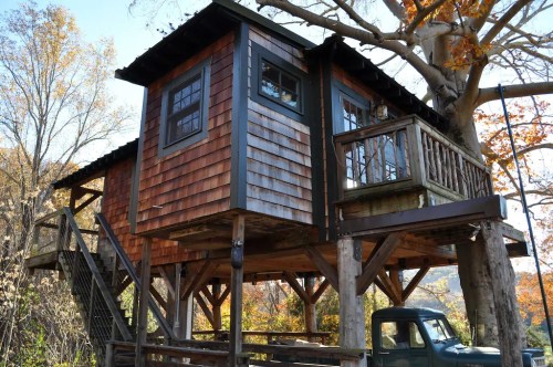 A rustic treehouse with wooden siding, elevated on stilts, surrounded by autumn foliage and a vintage truck below.