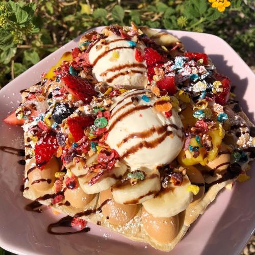 A colorful dessert featuring a waffle topped with bananas, strawberries, ice cream, and various toppings, drizzled with chocolate.