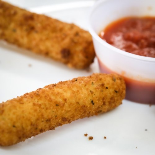Two golden-brown breaded cheese sticks on a plate, accompanied by a small cup of marinara sauce.