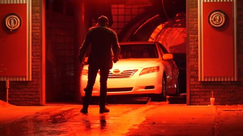 A person stands in front of a white car in a dimly lit garage, illuminated by red light.