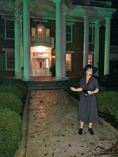 A woman in a black dress stands on a path in front of a large, illuminated house with green lighting and columns.