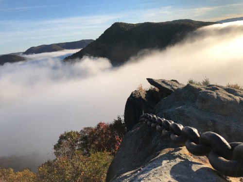 A chain rests on a rocky ledge overlooking a foggy valley and distant mountains under a clear blue sky.