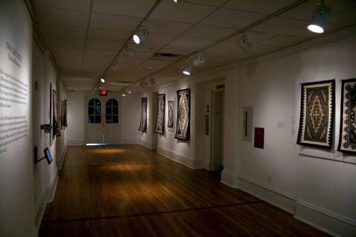 A gallery interior featuring walls adorned with framed textile art and wooden flooring, illuminated by overhead lights.