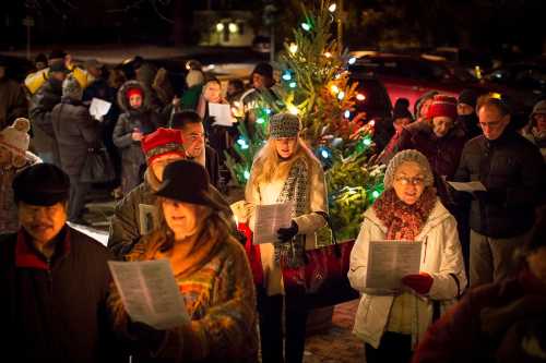 A group of people singing carols around a decorated Christmas tree at night, wearing winter clothing.