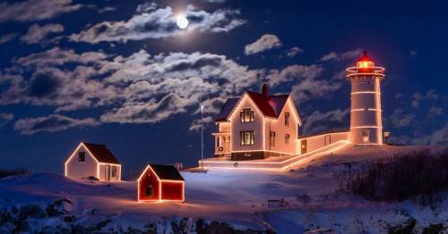 A beautifully lit lighthouse and house under a full moon, surrounded by snow and clouds in a serene night scene.