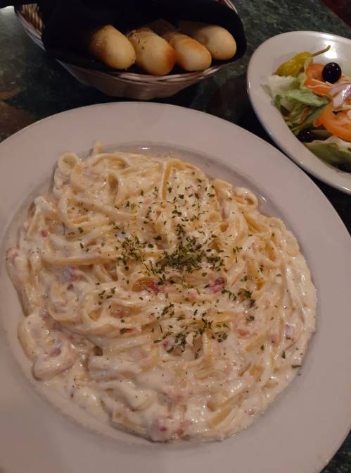 A plate of creamy fettuccine alfredo topped with parsley, accompanied by a side salad and breadsticks in a basket.
